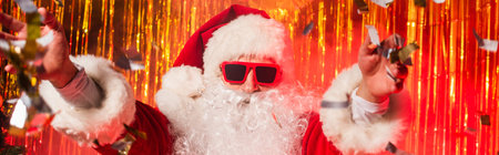 Santa claus in sunglasses and costume throwing confetti during party near tinsel, bannerの写真素材