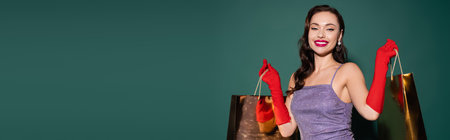 happy young woman in red gloves and purple dress holding shopping bags on green, bannerの写真素材