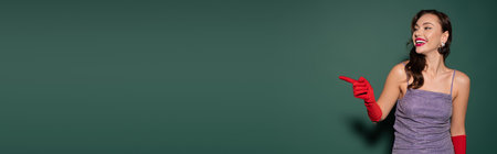 brunette young woman in red gloves and purple dress smiling while pointing with finger on green, bannerの写真素材