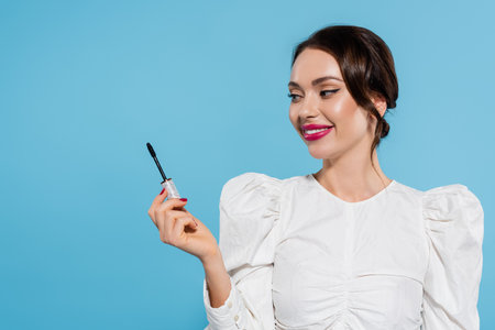 cheerful young woman in white blouse holding mascara brush isolated on blueの写真素材