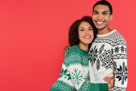young african american couple in sweaters with winter pattern embracing and smiling at camera isolated on redの写真素材