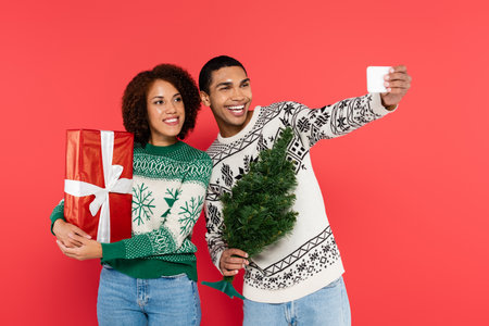 cheerful african american man with small christmas tree taking selfie with girlfriend holding...