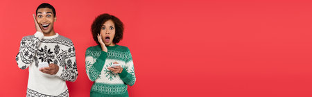 shocked african american woman in winter sweaters holding cellphones and looking at camera isolated...