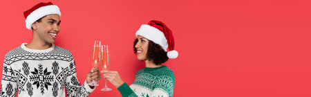 cheerful african american couple in santa hats and warm sweaters clinking champagne glasses isolated on red, bannerの写真素材