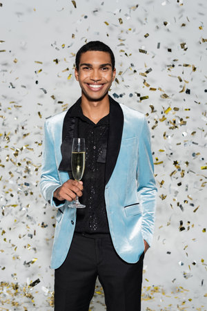 african american man in elegant clothes standing with champagne glass under confetti on grey...