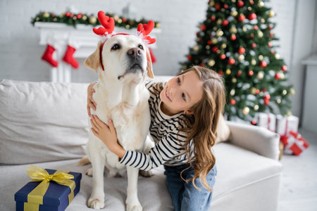 Kid hugging labrador with christmas headband near present on couch at homeの写真素材