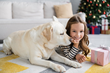 Labrador lying near smiling kid and christmas gift on carpet at homeの写真素材
