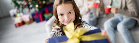 Smiling girl holding blurred christmas present and looking at camera at home, bannerの写真素材