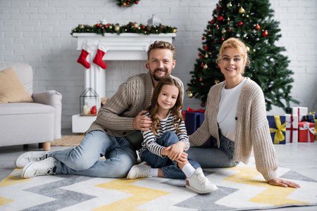 Father in knitted cardigan hugging daughter near wife during new year celebration at homeの写真素材