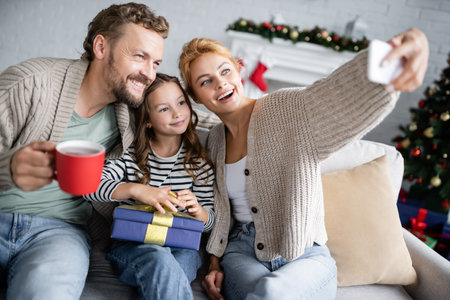 Woman taking selfie with family holding christmas gift and cup on couch at home