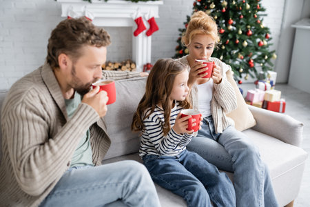 Woman looking at daughter with cup of cacao near husband on couch during new year at homeの写真素材