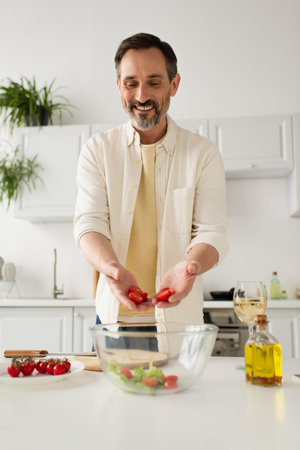 happy bearded man holding chopped cherry tomatoes near bowl with lettuce and glass  of white wineの写真素材
