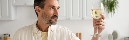 bearded man in white shirt looking at glass of white wine in kitchen, banner