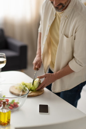 partial view of blurred man cutting fresh lettuce near mobile phone with blank screenの写真素材