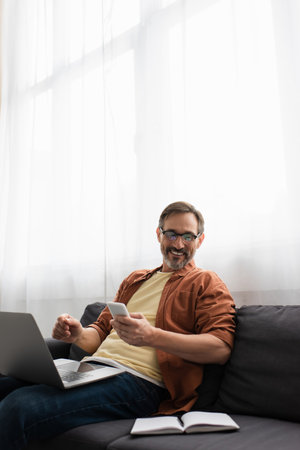 smiling bearded man holding smartphone and looking at empty notebook while sitting on sofa with...