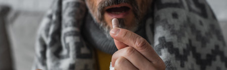 partial view of diseased man taking pill while sitting under warm blanket, banner