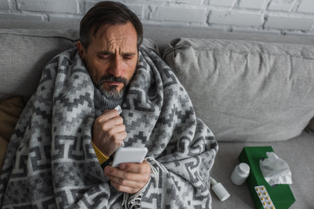 man covered with warm blanket looking at smartphone while sitting near containers with treatment and pack of paper napkinsの写真素材