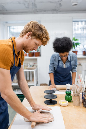 redhead man in apron shaping clay piece with rolling pin near african american woman during pottery...