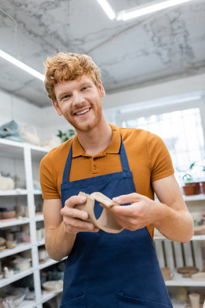 cheerful man with red hair standing in apron and modeling clay piece during pottery classの写真素材