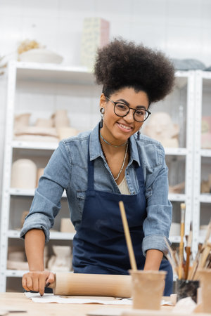 cheerful african american woman in eyeglasses and apron modeling clay with rolling pinの写真素材