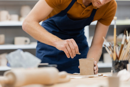 cropped view of bearded man in apron shaping clay piece into cup