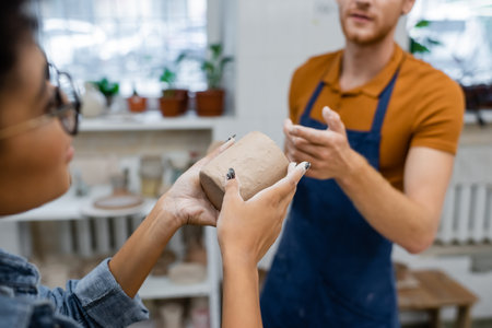 bearded man pointing with finger at clay cup in hands of african american woman in pottery studioの写真素材