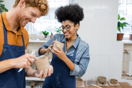 cheerful african american woman in eyeglasses molding clay cup with happy redhead boyfriend in...