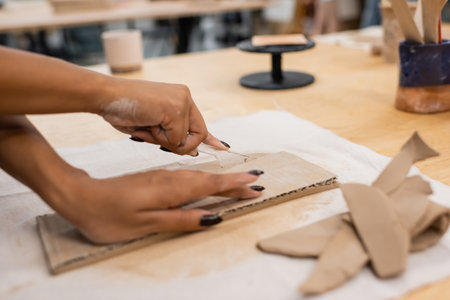 partial view of african american woman cutting clay with knife while handcrafting in pottery...