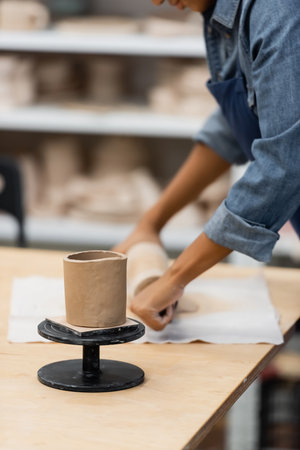 clay cup on stand near african american woman handcrafting with rolling pin during pottery class