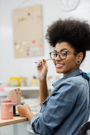 Smiling african american woman in eyeglasses holding paintbrush near clay sculpture and paint in pottery studioの写真素材