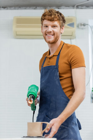 Smiling redhead craftsman in apron holding heat gun near blurred ceramic cup in pottery workshopの写真素材
