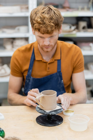 Redhead artisan in apron looking at ceramic cup in pottery workshopの写真素材