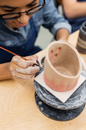 Young african american woman in eyeglasses painting on blurred clay product in pottery studioの写真素材