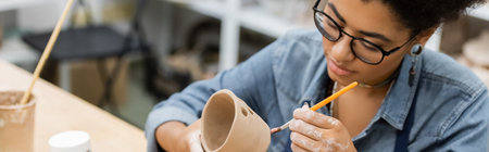Young african american craftswoman in eyeglasses painting clay product in pottery workshop, banner