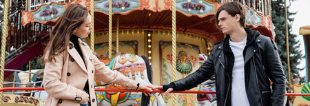 young couple standing near carousel and touching hands in amusement park, bannerの写真素材