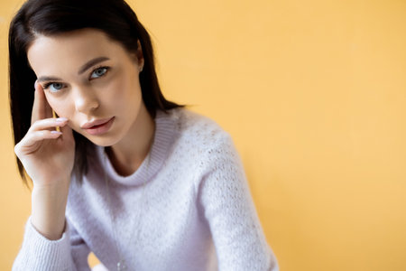 brunette woman with natural makeup wearing cozy sweater and looking at camera isolated on yellowの写真素材