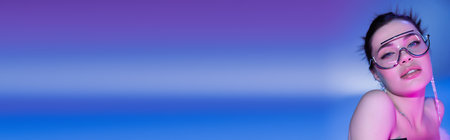 portrait of sensual brunette woman in transparent sunglasses looking at camera on blue and purple...