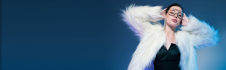 brunette woman in transparent sunglasses and white furry jacket standing with hands behind head on blue background, bannerの写真素材