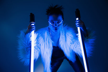 Trendy young woman in furry jacket and sunglasses holding lamps on navy blue background