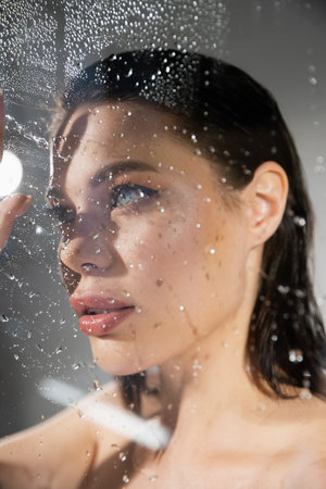 Young brunette woman standing near wet glass on grey background