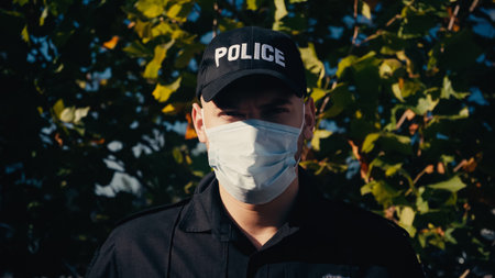 portrait of young policeman in uniform and medical mask standing near treeの写真素材