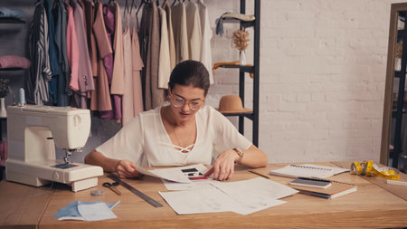 Seamstress holding sketches near sewing machine and notebooks in atelierの写真素材