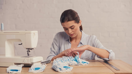 Brunette seamstress counting medical masks near sewing machine and ruler in atelier