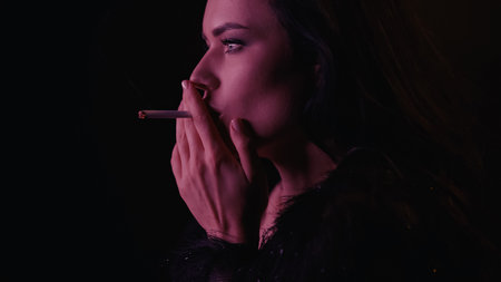 Young woman smoking cigarette and looking away isolated on black with lighting
