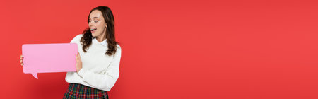 Pretty young woman in sweater holding speech bubble isolated on red, bannerの写真素材