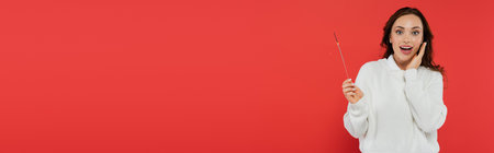 Excited woman in cozy sweater holding sparkler and looking at camera isolated on red, banner