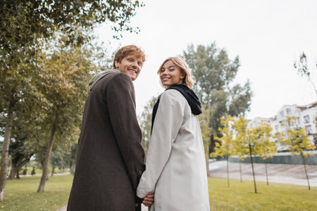 low angle view of joyful blonde woman and cheerful redhead man holding hands while strolling in parkの写真素材
