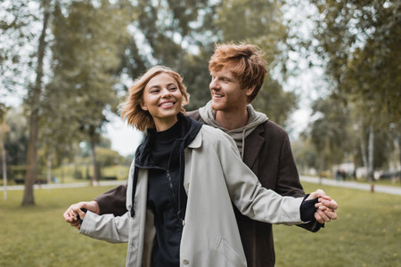 positive young man in coat holding hands with cheerful blonde girlfriend in autumnal parkの写真素材