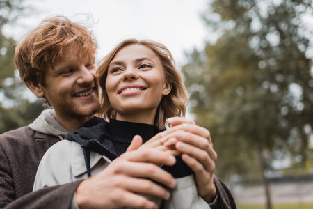 low angle view of caring young man in coat warming up hands of cheerful girlfriend in autumnal park