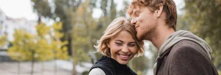 happy redhead man kissing head of blonde girlfriend smiling in park, banner
